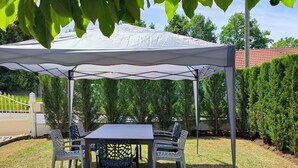 Un espacio cubierto al aire libre con mesa y sillas, rodeado de un seto y un edificio al fondo.