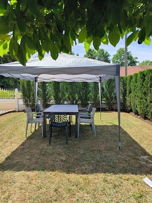 A covered outdoor dining area with a table and chairs, surrounded by a hedge and a building in the background.