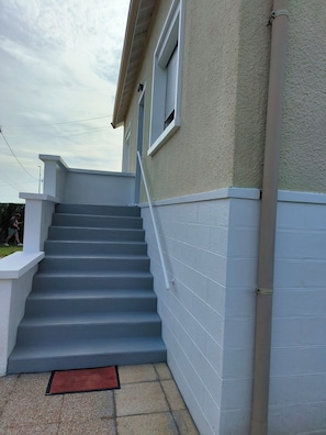 Un escalier extĂ©rieur mĂšne Ă un bĂątiment dont la façade est texturĂ©e et qui possĂšde une fenĂȘtre avec des stores fermĂ©s.