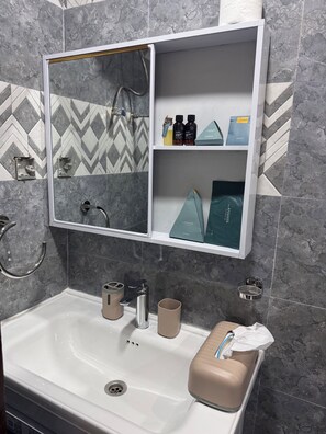 A bathroom with a white sink, a mirror with a shelf above it, and a towel dispenser. - Chic, modern apartment in GB, 3 min from town center. (Kinshasa)