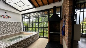 A spa area with a glass-enclosed hot tub, a brick fireplace, and large windows offering a view of greenery. - Country House in Quindío Casa Victoria (Calarcá)