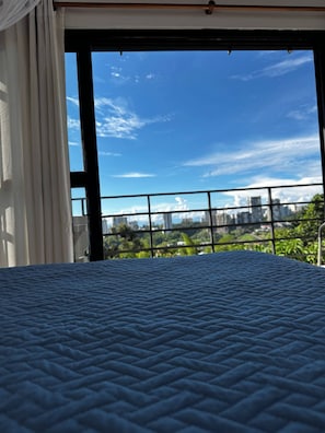 Ein Zimmer mit Bett, Blick auf eine Stadtlandschaft durch ein Fenster und ein Balkon mit Geländer.