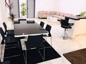 Ein modernes Wohnzimmer mit einem beigen Sofa, einem Esstisch mit Stühlen, einem Couchtisch und einer kleinen Küchenzeile mit Theke und Stuhl.