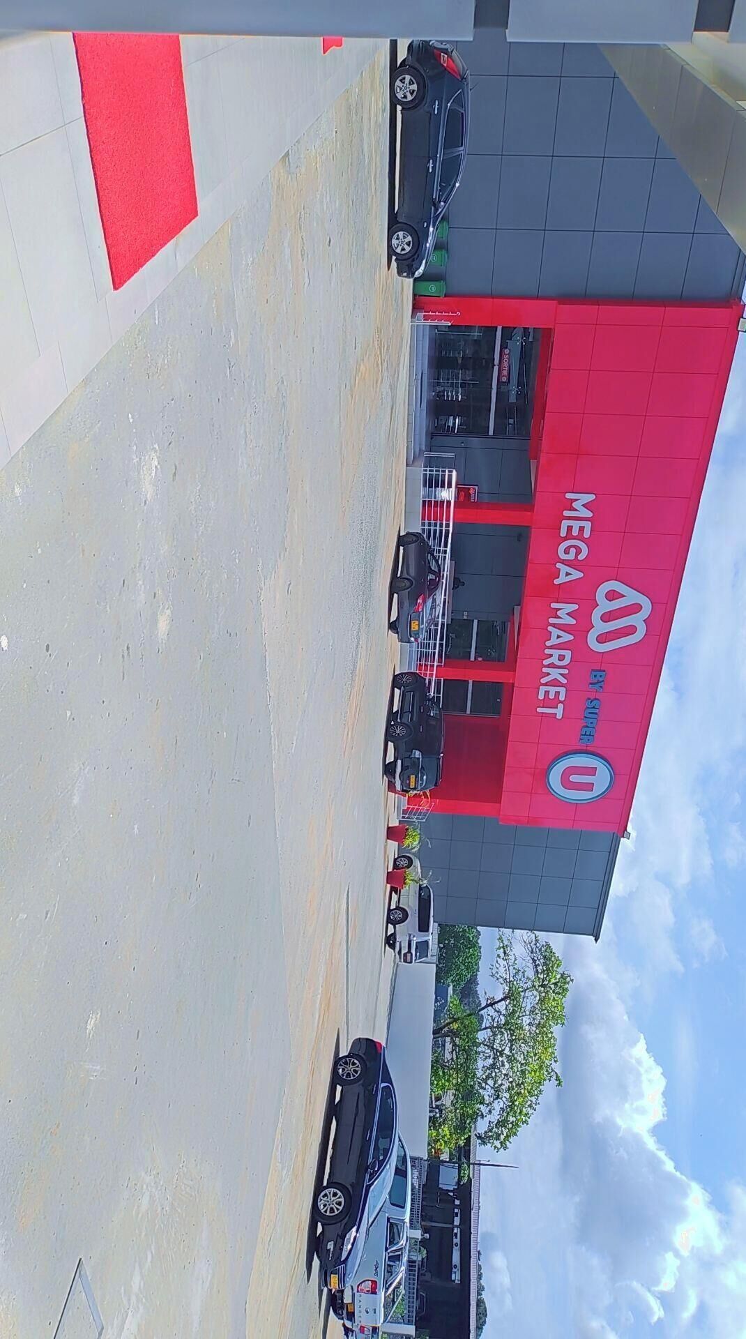 Un estacionamiento con varios coches, un edificio de fachada roja y gris, y un cielo despejado.