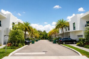 Une vue d’une rue dans un quartier résidentiel, avec des immeubles modernes aux façades blanches, des voitures garées et des palmiers.