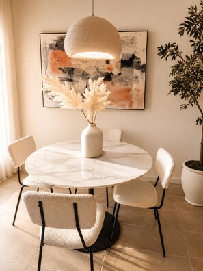 Une table de salle à manger ronde, accompagnée de quatre chaises, un vase contenant de l’herbe des pampas, une grande peinture abstraite et une plante en pot.