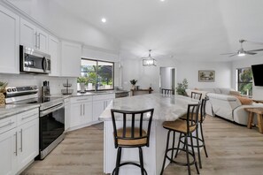 Eine moderne Küche mit weißen Schränken, Edelstahlgeräten, einer großen Kochinsel mit Barhocker und einem Essbereich mit Sofa und Tisch.
