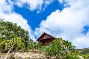 A wooden house with a balcony, surrounded by tropical vegetation and a stone wall. - Cozzy Pini Sentana Village Nusa Penida  (Klungkung)