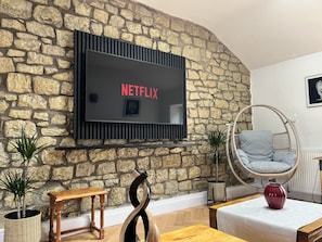 Ein Wohnzimmer mit Steinwand, einem an der Wand befestigten Fernseher, einem Holztisch, einem Korbstuhl und Topfpflanzen.