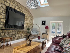 Ein Wohnzimmer mit Steinwand, Flachbildfernseher, Korbsessel, Couchtisch aus Holz, Ledersofa und einer Kronleuchter.