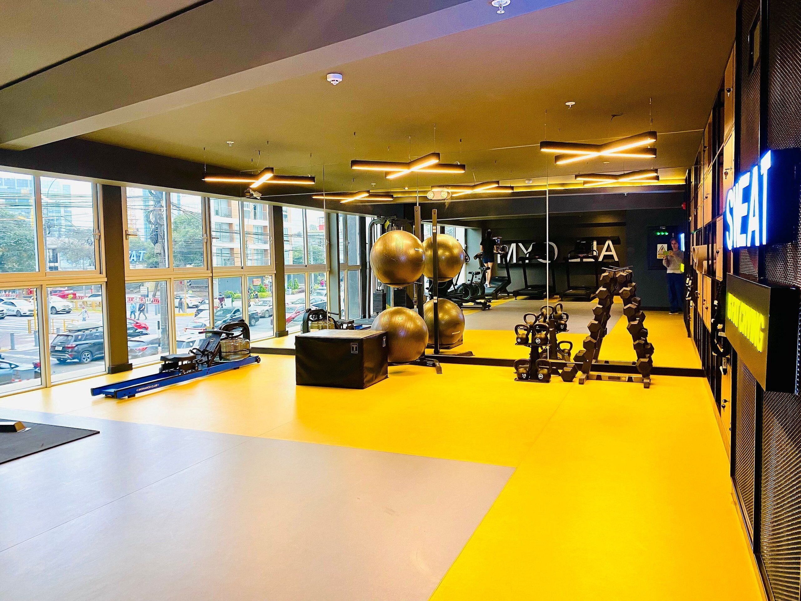 Une salle de sport équipée de matériel d’exercice, dotée de grandes fenêtres et d’un sol jaune.