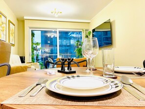 Mesa de comedor dispuesta con platos, cubiertos y copas de vino, con vistas a un paisaje urbano a través de amplios ventanales.