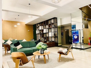 Um hall moderno com um sofá verde, cadeiras de madeira, uma estante de livros e um balcão de recepção.
