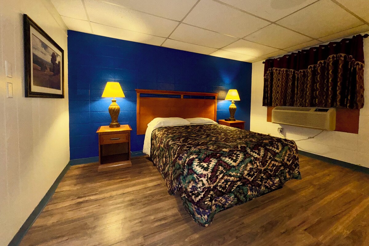 Una habitación de hotel con cama de madera, dos lámparas de noche, un cuadro enmarcado en la pared y una ventana con cortinas.
