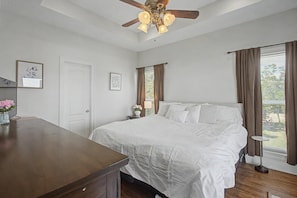 Ein Schlafzimmer mit einem Bett, einem Deckenventilator, einem Nachttisch mit Lampe, einem Fenster mit Vorhängen und einer Tür.