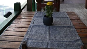 Una terraza de madera con mesas y sillas, una mesita con un jarrón de flores y vistas a un cuerpo de agua con barcos.