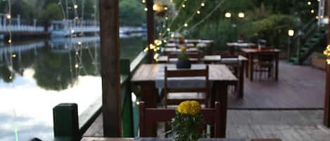 Una terraza de madera con mesas y sillas, una mesita con un jarrón de flores y vistas a un cuerpo de agua con barcos.