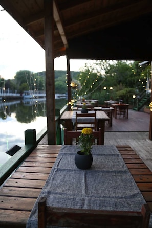 A wooden deck with tables and chairs, a small table with a vase of flowers, and a view of a body of water with boats. - Mell's River (Sile)