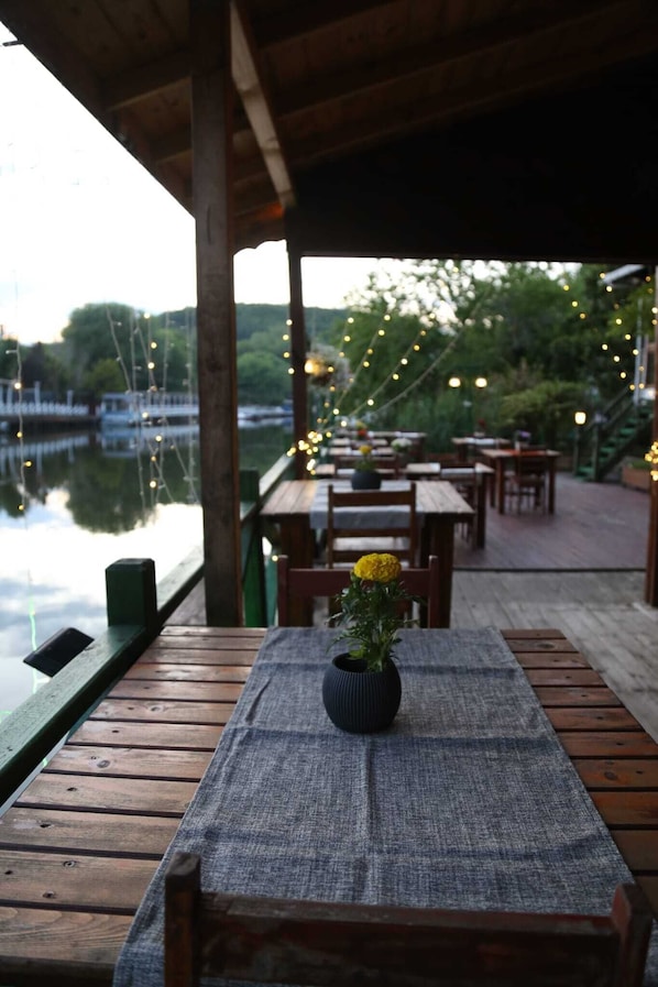 A wooden deck with tables and chairs, a small table with a vase of flowers, and a view of a body of water with boats.