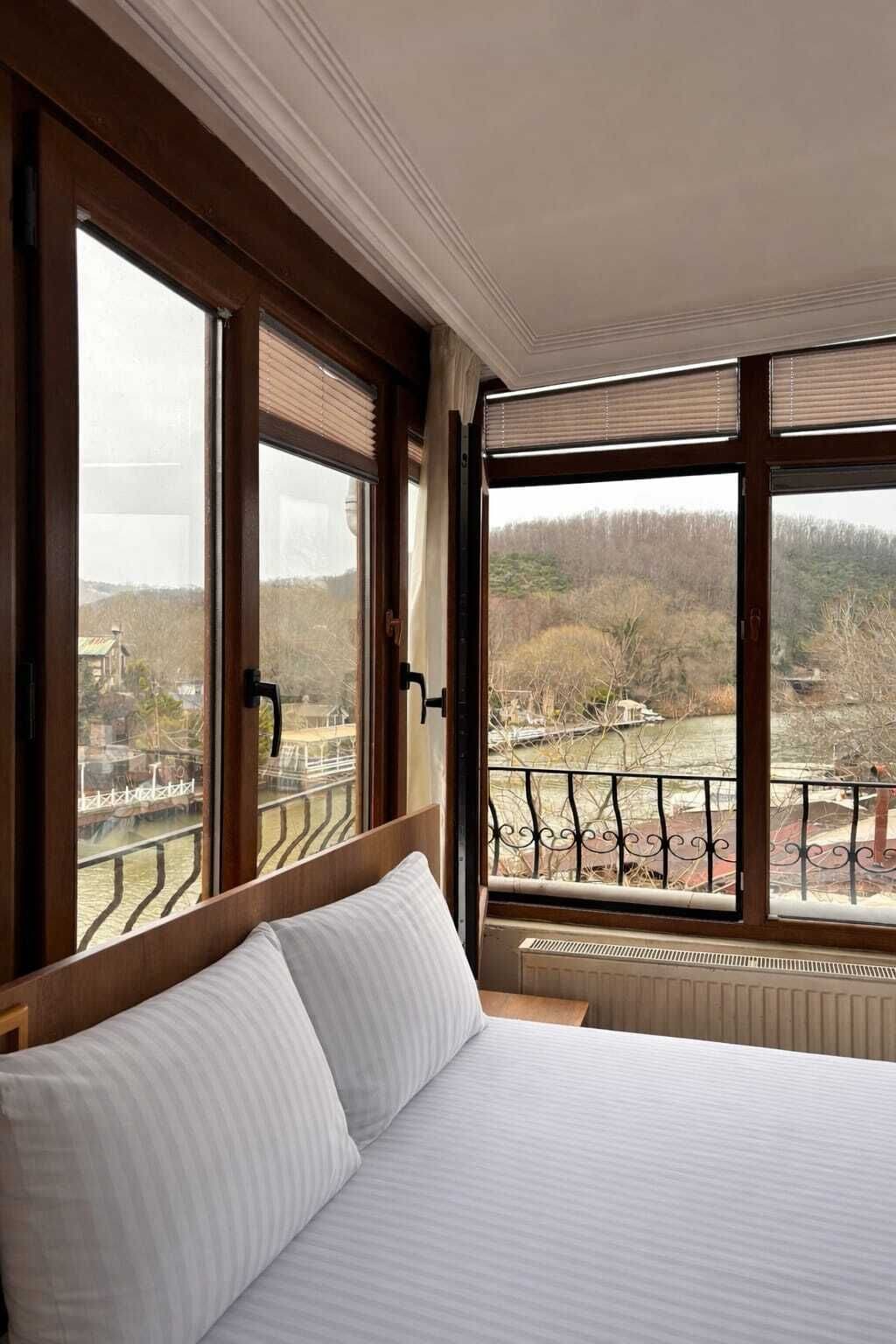 A bedroom with a large bed, two windows with blinds, and a view of a river and hills.