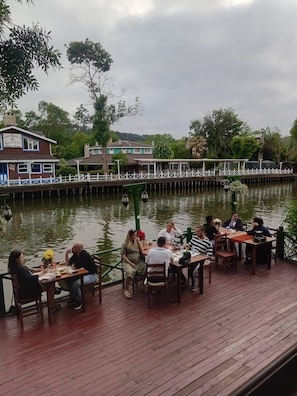 A wooden deck with tables and chairs, overlooking a body of water, with buildings and trees in the background. - Mell's River (Sile)