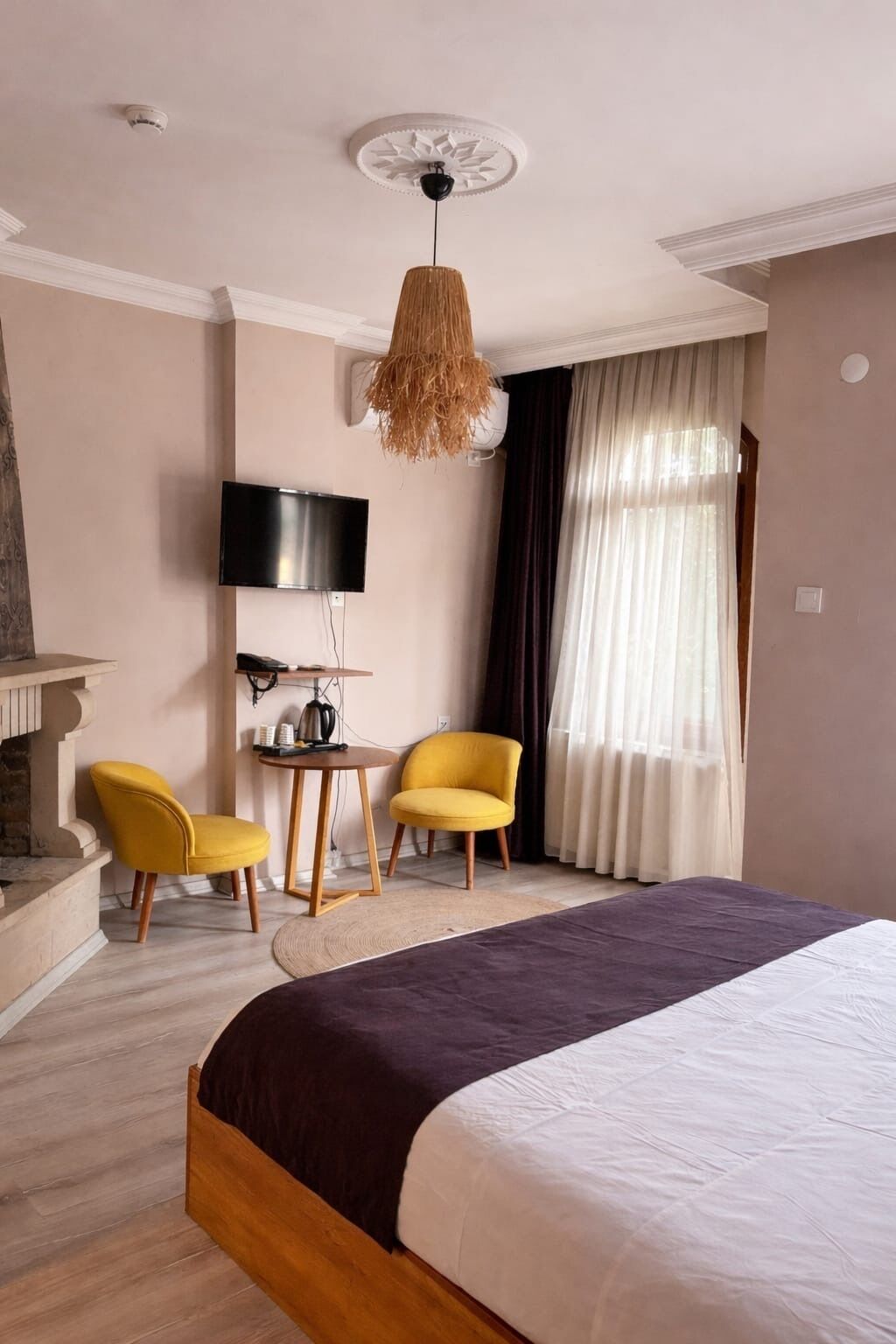 A hotel room with a bed, two yellow chairs, a TV, a fireplace, and a ceiling fan.
