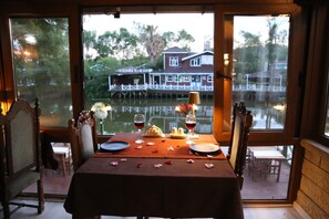 A dining table set with wine glasses, plates, and a flower centerpiece, overlooking a houseboat and water. - Mell's River (sile)