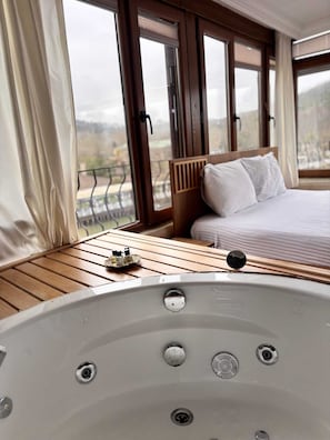 A bedroom with a large window, a wooden headboard, a bed with white bedding, and a wooden table with a bottle and a glass.
