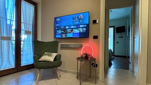 A living room with a television mounted on the wall, a green armchair, a small table with a red light, and a sliding glass door. - Mazzini28, A1 Caserta North, Royal Palace, concerts, Naples, coast, southern Italy (Casapulla)
