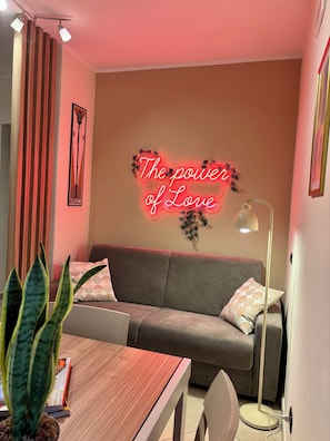 Ein gemütlicher Wohnbereich mit einer Couch, einem Tisch mit Pflanze, einer Lampe und einem Neon-Schild an der Wand.
