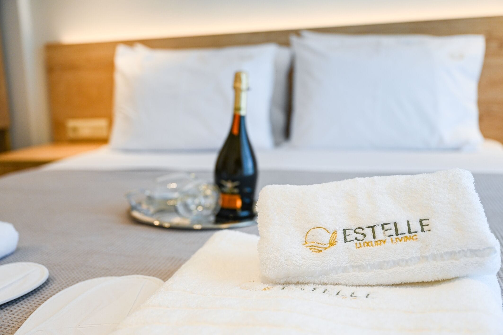 Ein ordentlich bezogenes Bett mit einer Champagnerflasche, einem Tablett mit Gläsern und einem Handtuch mit dem Aufdruck 'ESTELLE LUXURY LIVING'.