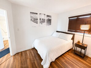 Een slaapkamer met een houten vloer, een bed met wit bedlinnen, een nachtkastje met een lamp, en drie zwart-wit foto’s aan de muur.