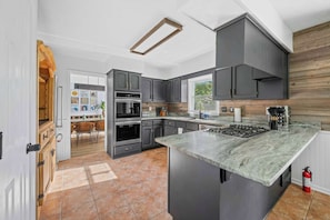 A modern kitchen with grey cabinets, a central island, and a dining area visible through an open door.