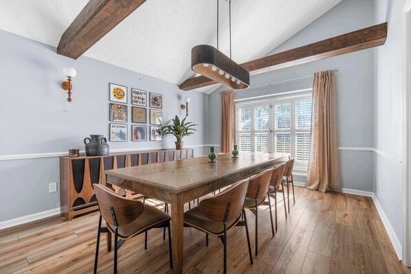 A dining room with a wooden table and chairs, a ceiling fan, and a window with curtains.