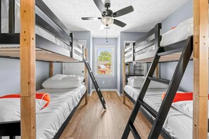 A bunk bed room with a ceiling fan, wooden floor, and a window with a view of trees.