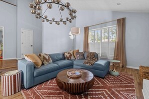 A living room with a sectional sofa, a round coffee table, a unique chandelier, and a large window with curtains. - Spacious Retreat w/ Hot Tub & Games | Sleeps 16 (Columbus)