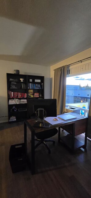 Una habitación con un escritorio de madera, una silla de oficina negra, una estantería y un ventanal con cortinas.