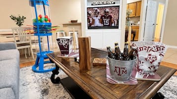 Un salon avec une table basse en bois, une machine à bonbons bleue, un téléviseur diffusant un match de football, et une cuisine en arrière-plan.