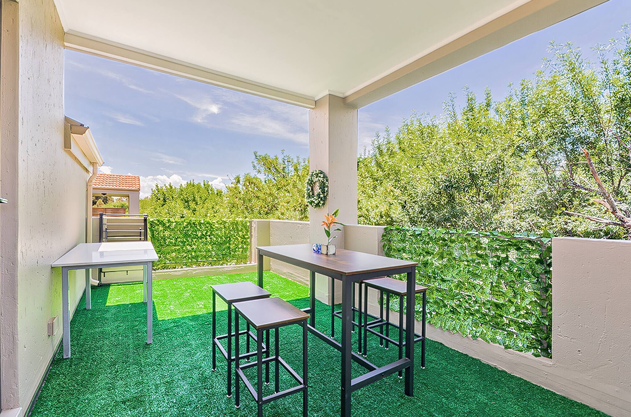 An outdoor patio with a table and chairs, a small table with a vase, and a view of a garden.