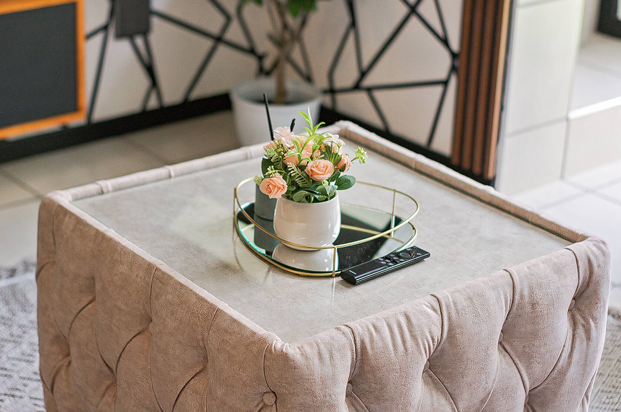 A beige tufted ottoman with a glass coffee table on top, a white vase with flowers, and a remote control.