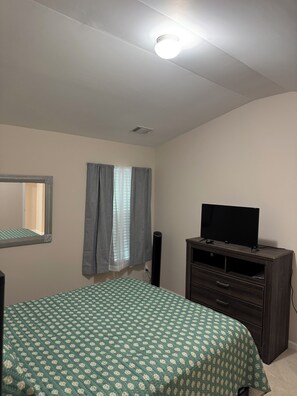 Ein Schlafzimmer mit einem Bett, einem Schminktisch, einem Fernseher, einem Spiegel und einem Fenster mit Vorhängen.