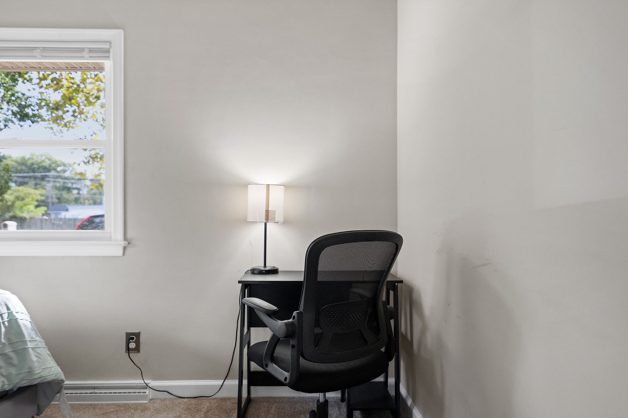 A room with a desk, chair, lamp, and a window with a view of trees and a car.