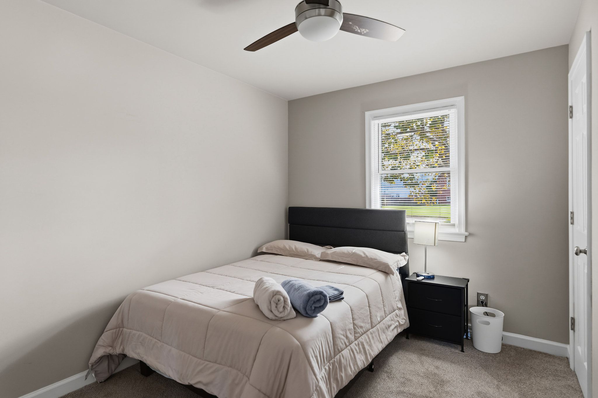 A bedroom with a bed, a nightstand, a lamp, a ceiling fan, and a window with blinds.