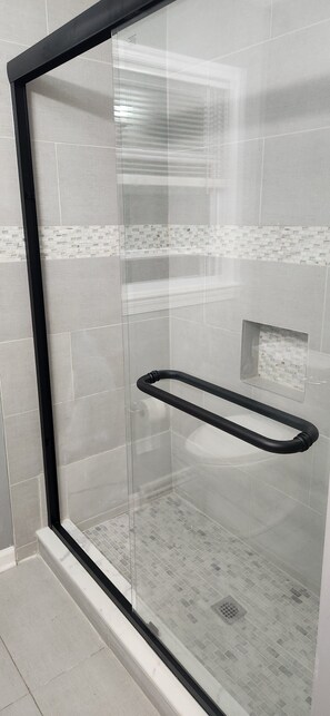 Une douche fermée par des parois en verre, dotée d’une poignée noire, de murs carrelés et d’une petite fenêtre.