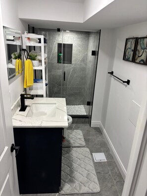 A bathroom with a white sink, a glass shower enclosure, a towel rack with yellow towels, and a small shelf with toiletries. - Luxury Guest Suite (Thorold)