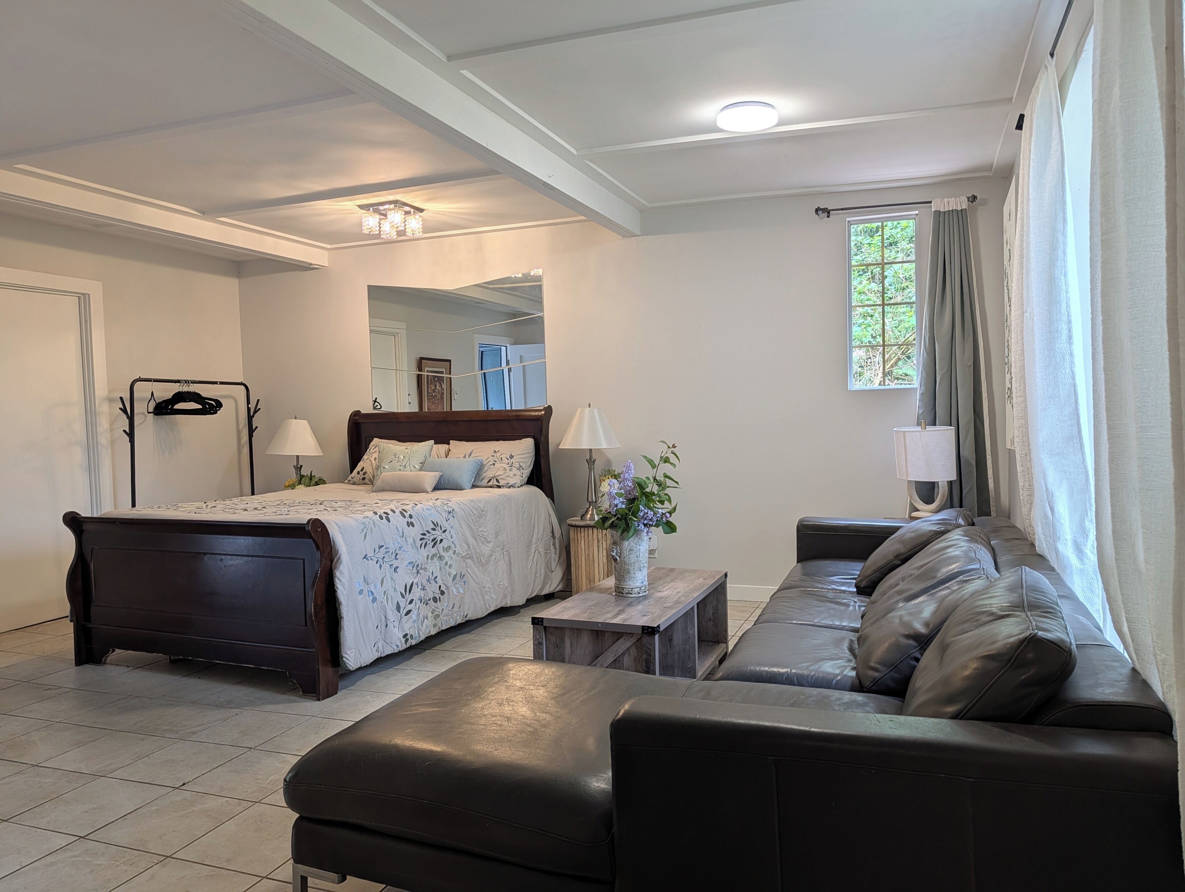 A bedroom with a bed, a sofa, a small table, a mirror, and a window with curtains.