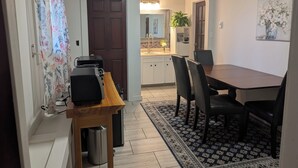 A dining area with a table and chairs, a kitchen in the background, and a door leading to another room.