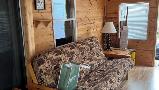 A wooden cabin interior with a sofa, a table, a lamp, and a window with blinds.