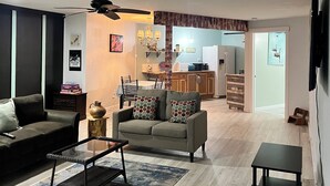Ein Wohnzimmer mit einer Couch, einem Couchtisch und einem Fernseher. Im Hintergrund ist eine Küche mit einer Essecke zu sehen.