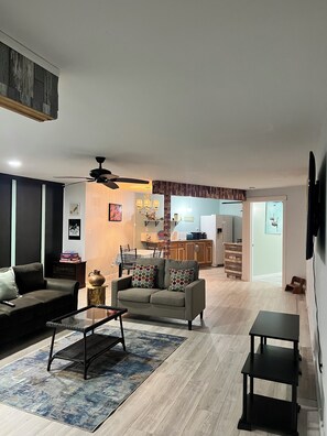 Una sala de estar con sofá, mesa de centro y televisión. Se ve una cocina al fondo con comedor.